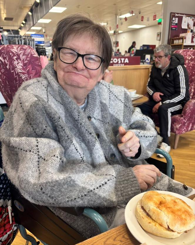 Elderly person in a café smiling and pointing while seated with a sandwich.