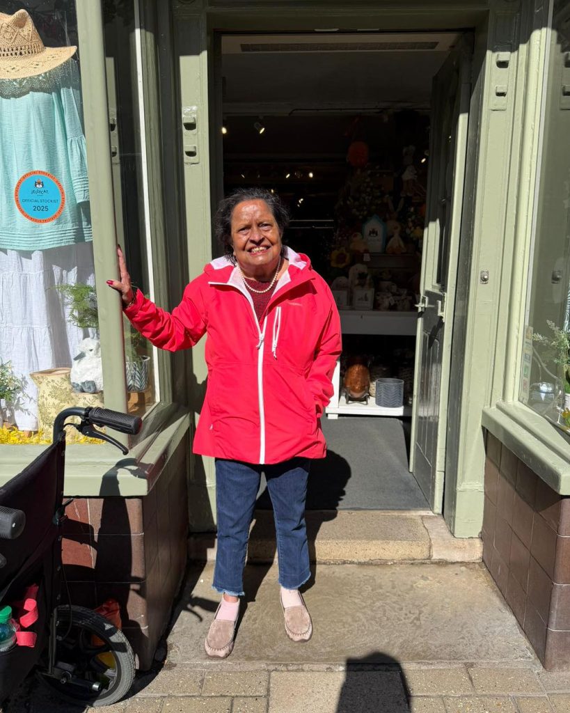 Smiling woman in a bright red jacket standing in a sunny doorway of a small shop.