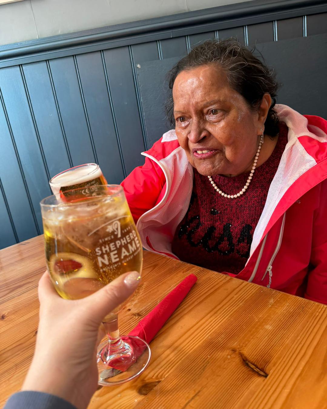 Two people clinking drinks at a wooden table, with a woman in a red jacket smiling.
