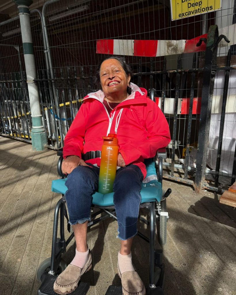 Smiling elderly woman in a red jacket sitting in a wheelchair outdoors near construction barriers.