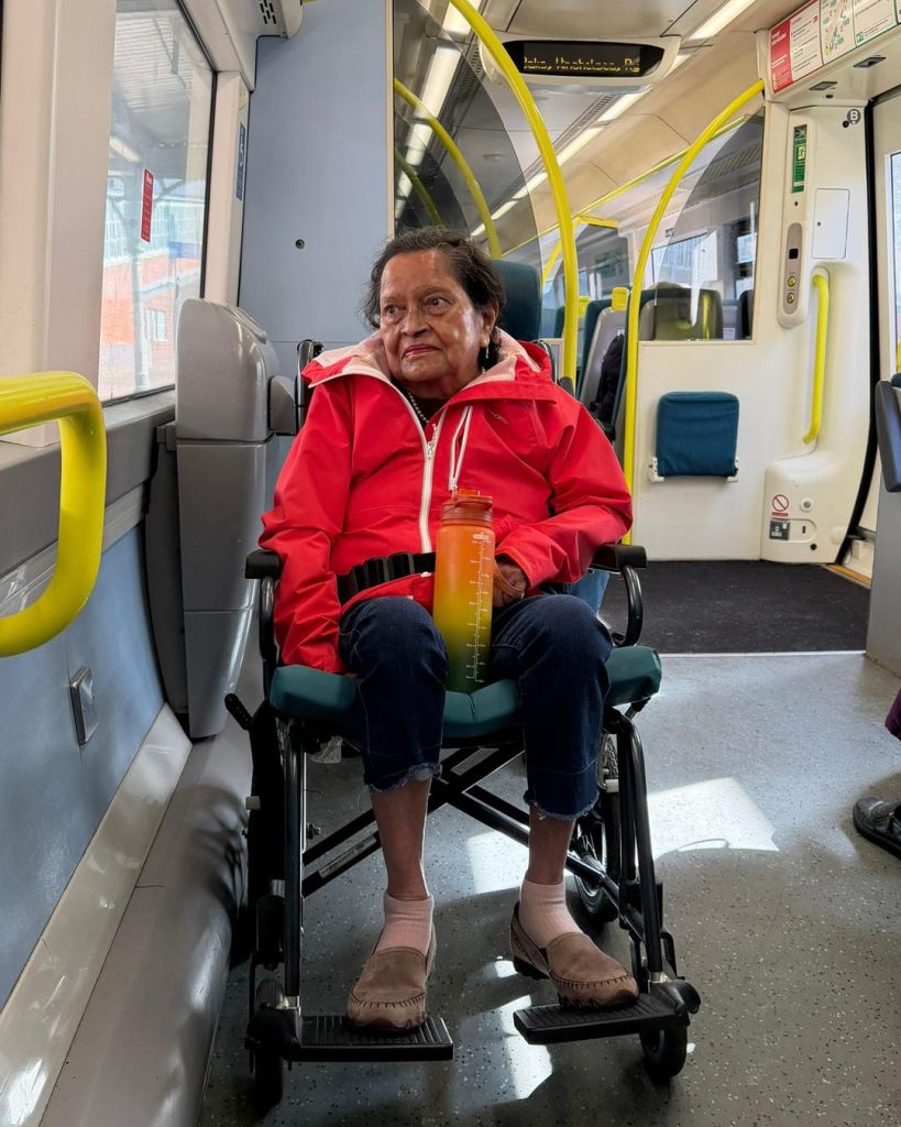 Elderly woman in a red jacket sitting in a wheelchair on a train, holding a water bottle.