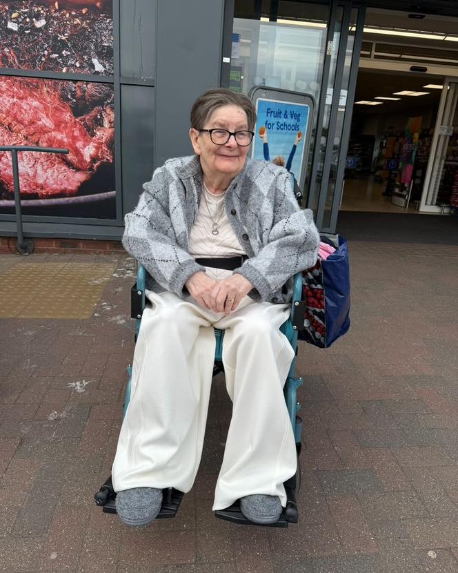 Elderly woman sitting in a wheelchair outside a store, wearing a grey cardigan.