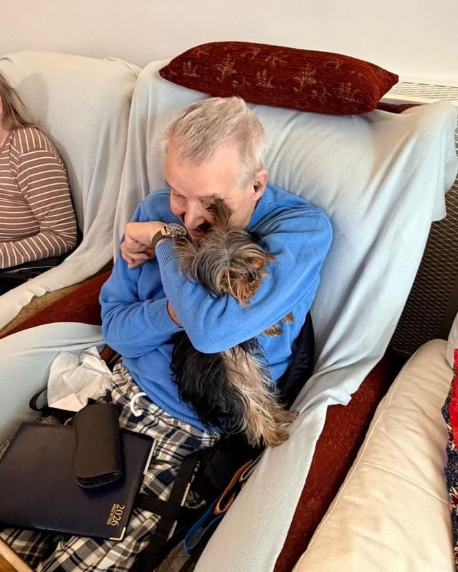 Older man cuddling a small dog closely while seated indoors.