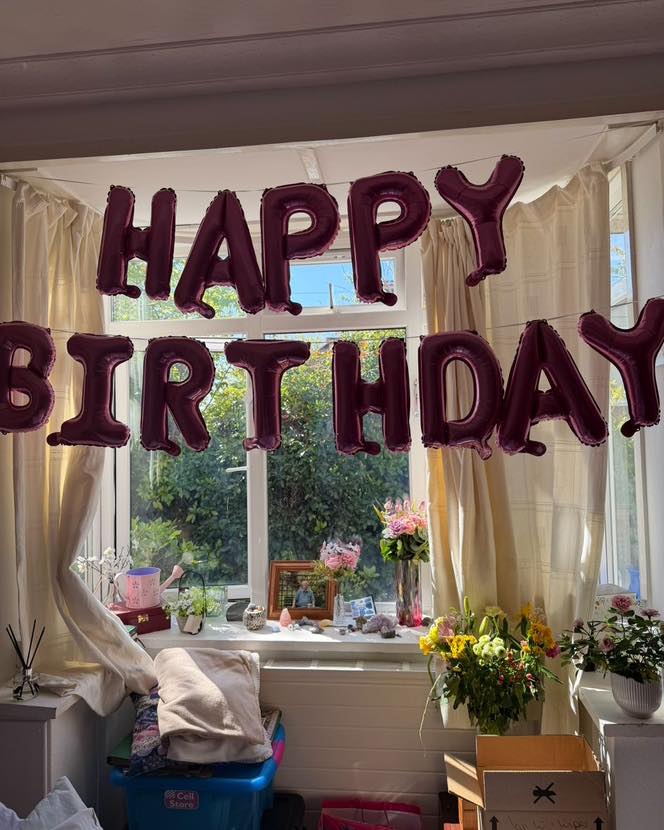 Pink ‘Happy Birthday’ balloon banner above a window with flowers and soft home decor.