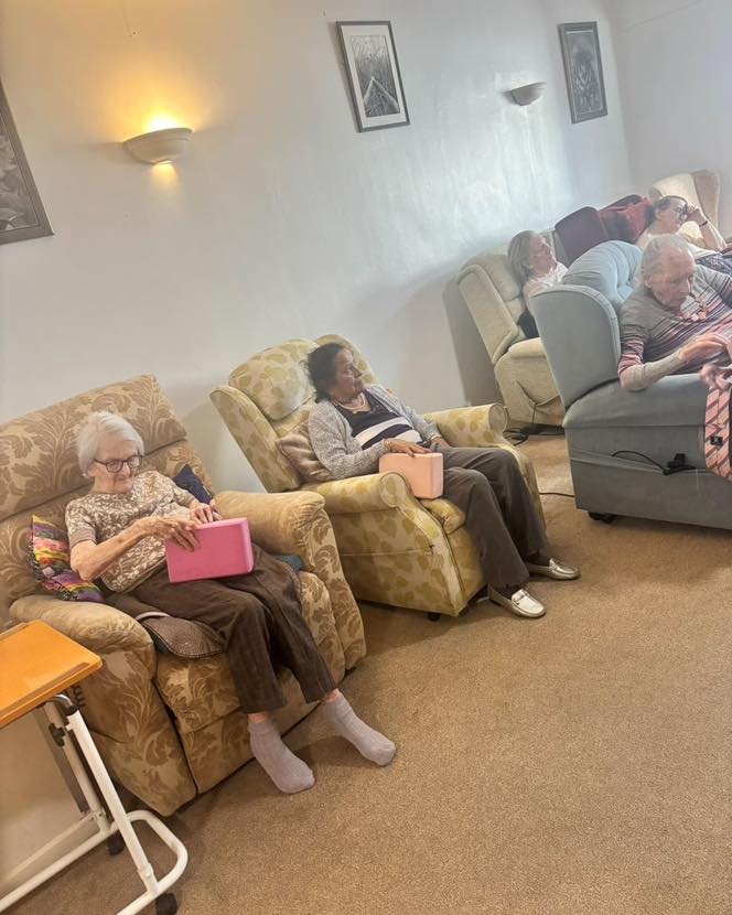 Elderly residents seated in a lounge holding pink foam blocks during a group activity.