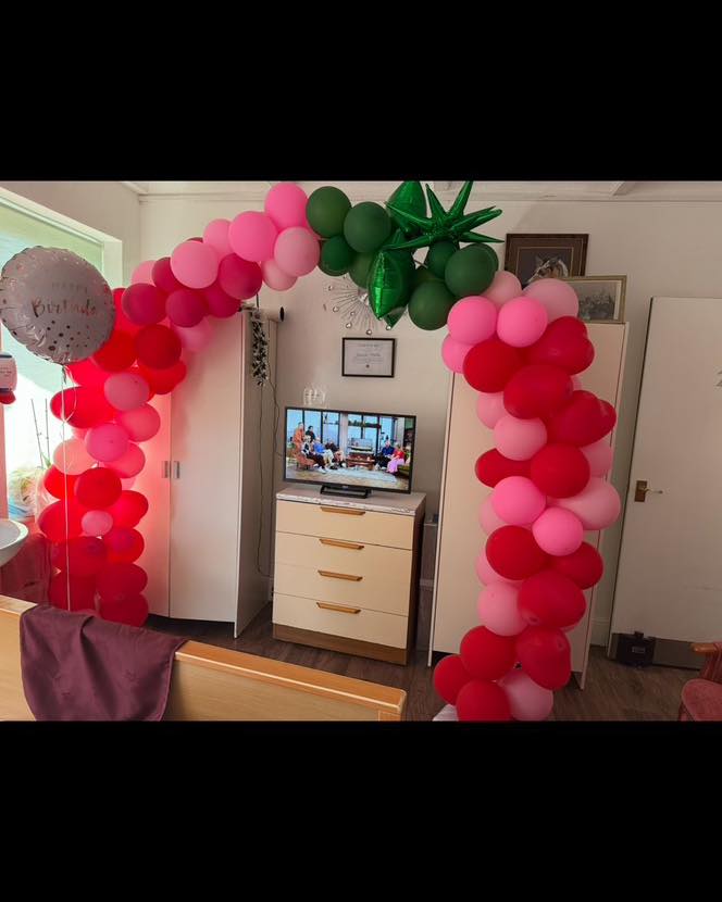Red, pink, and green balloon arch set up indoors around a TV cabinet.