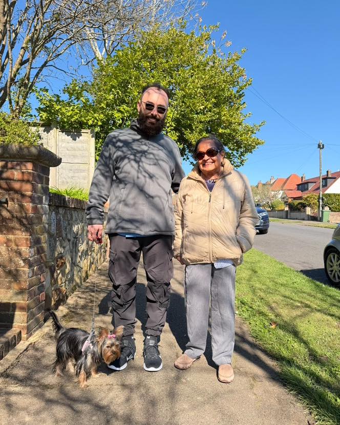 Man and woman walking a small dog together on a sunny sidewalk.