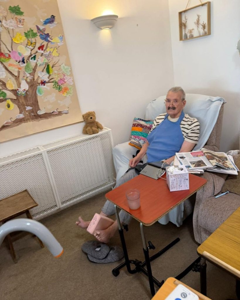 Elderly man sitting comfortably in a care home lounge beside a colorful wall display and small table.