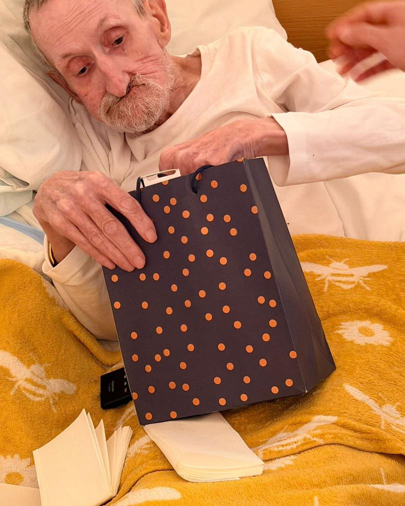 Older man in bed reaching into a gift bag during a bedside celebration.