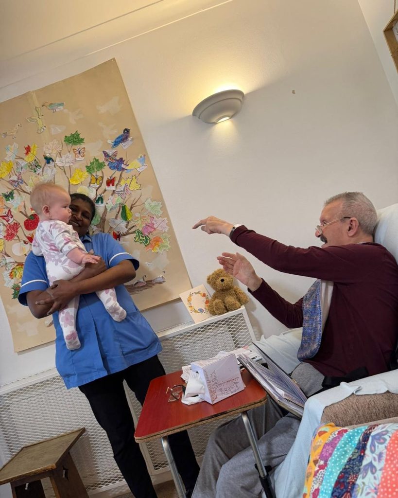 Care worker holding a baby while an elderly resident smiles and reaches out to interact.