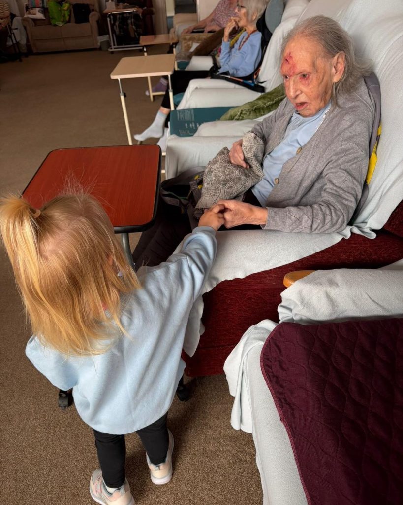 Young child holding hands with an elderly resident seated in a care home lounge.