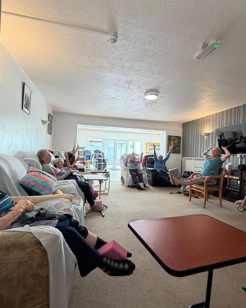 Care home residents taking part in a seated exercise session led by an activity coordinator in a bright communal room.