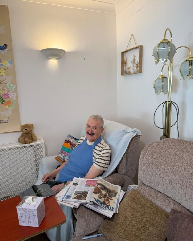 Smiling older man relaxing in an armchair with newspapers and personal belongings nearby in a cozy care setting.