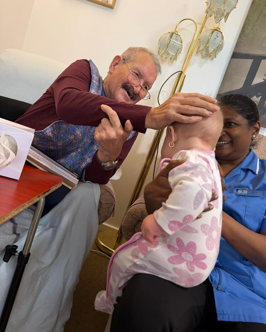 Elderly resident gently touching a baby's head while a caregiver supports the child.