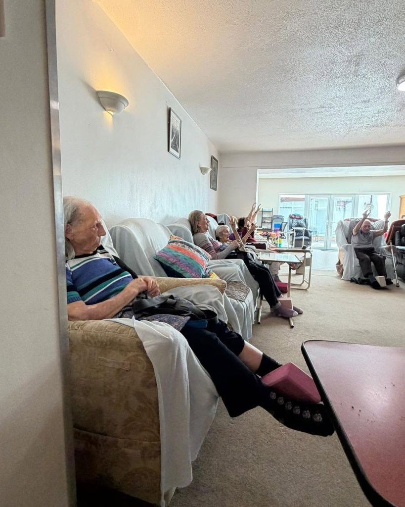 Older adults seated together in a care home lounge participating in a gentle group activity.