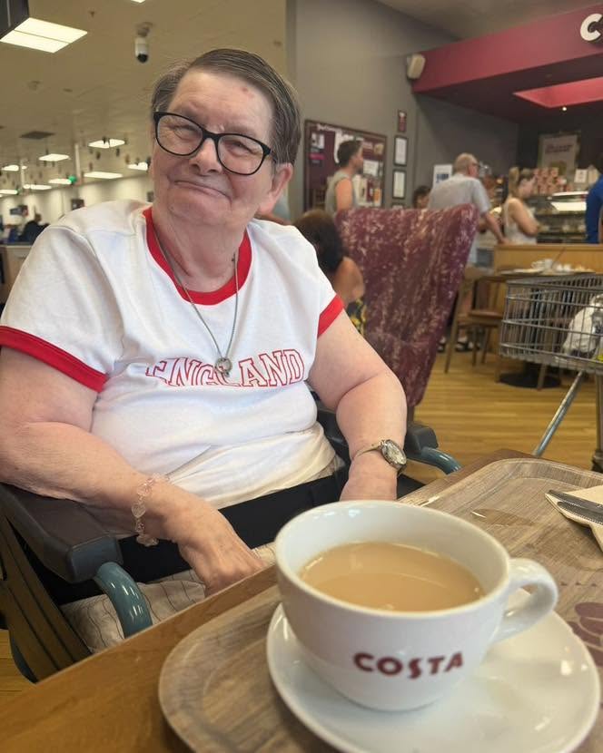 Smiling elderly woman sitting in a wheelchair at a Costa Coffee café with a cup of tea on the table.