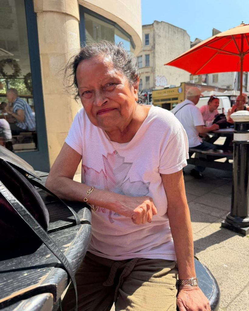 Elderly woman sitting on an outdoor bench in the sunshine, smiling at the camera.